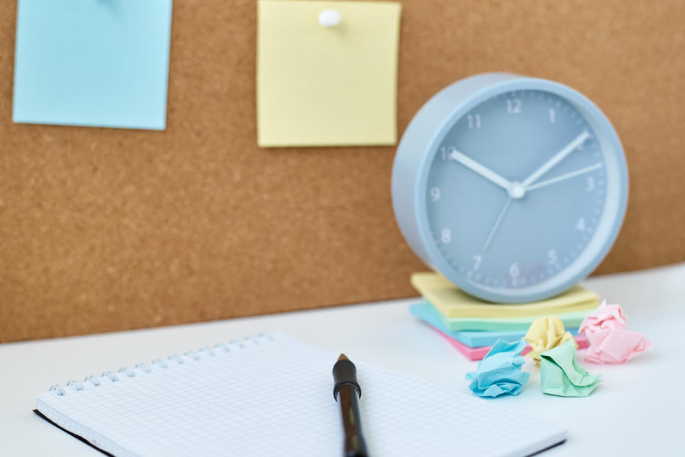 Clock next to sticky notes and crumpled paper on a desk, with a notebook and pen in front and a corkboard with sticky notes behind