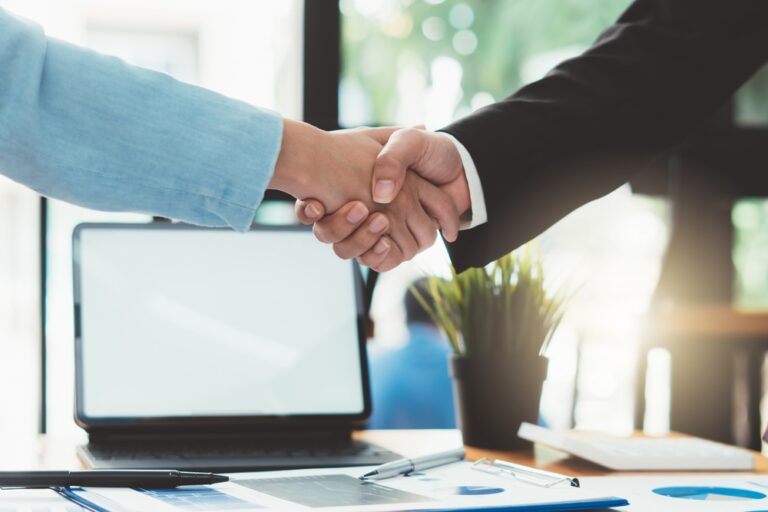 Two people shaking hands over a desk with a laptop and documents