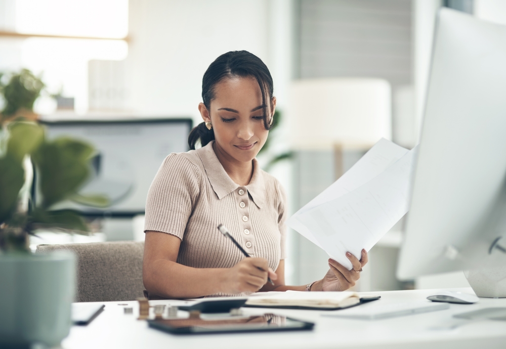 Woman reviewing a document while writing notes at a desk with a computer monitor