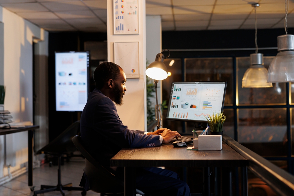 Man working at a desk with a monitor displaying charts and graphs, and a screen with similar data visible in the background