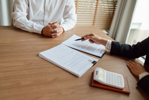 Man with clasped hands sitting across from a person pointing a pen at a document on a clipboard