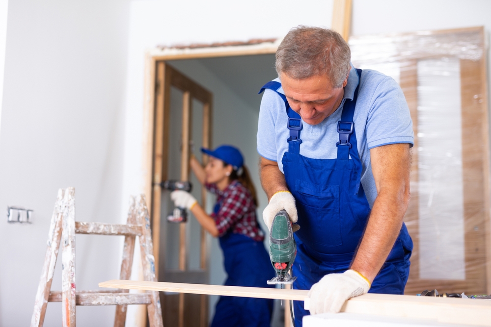 Man cutting a wooden board with a jigsaw while a woman drills a doorframe in the background