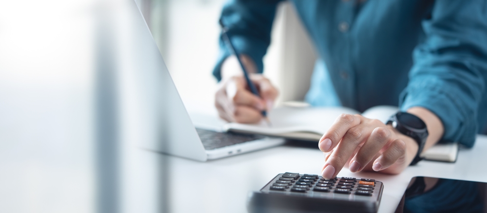 Person using a calculator while writing in a notebook next to a laptop