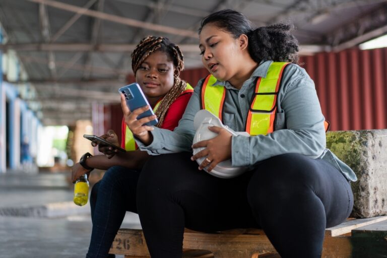 Two women wearing safety vests looking at a phone together while seated in a warehouse