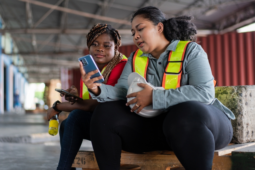 Two women wearing safety vests looking at a phone together while seated in a warehouse