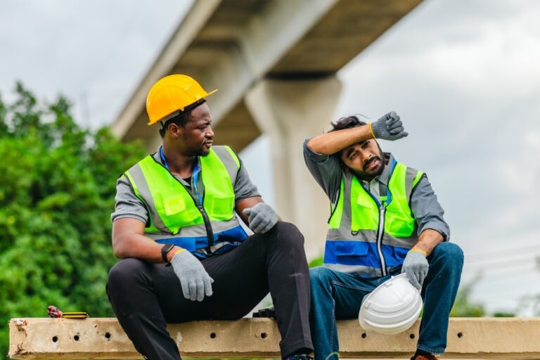 Two construction workers sitting on a concrete beam under an overpass, one wiping sweat from his brow