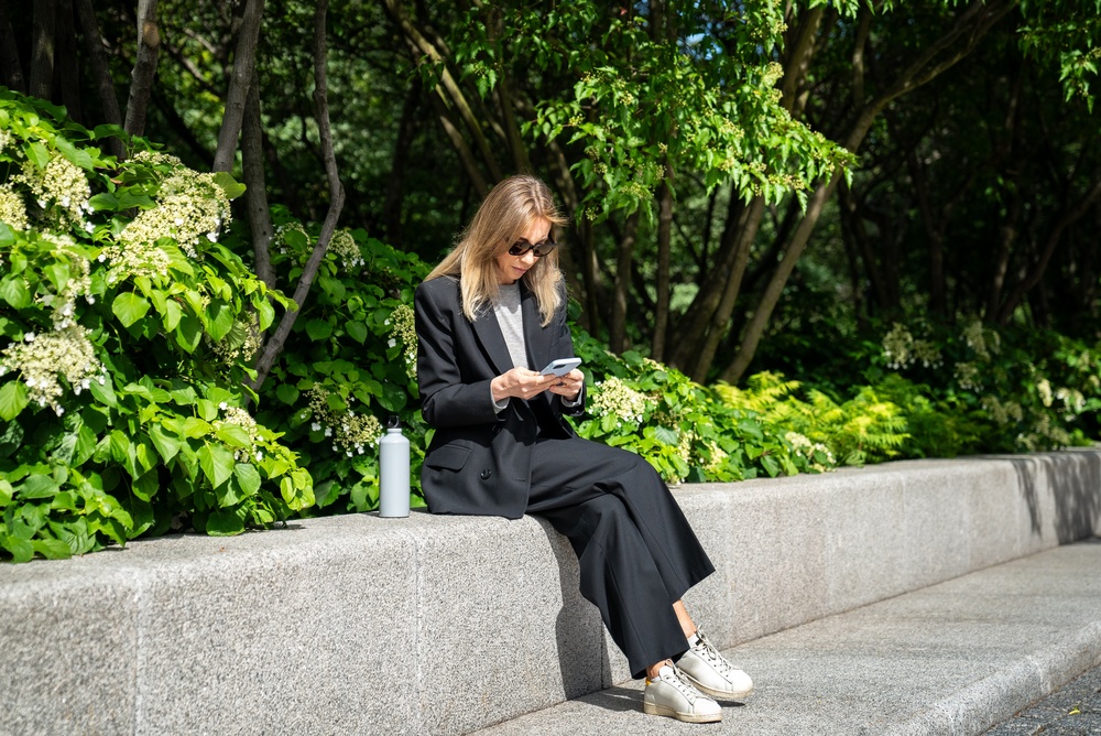 Woman sitting on a stone ledge in a park, looking at her phone
