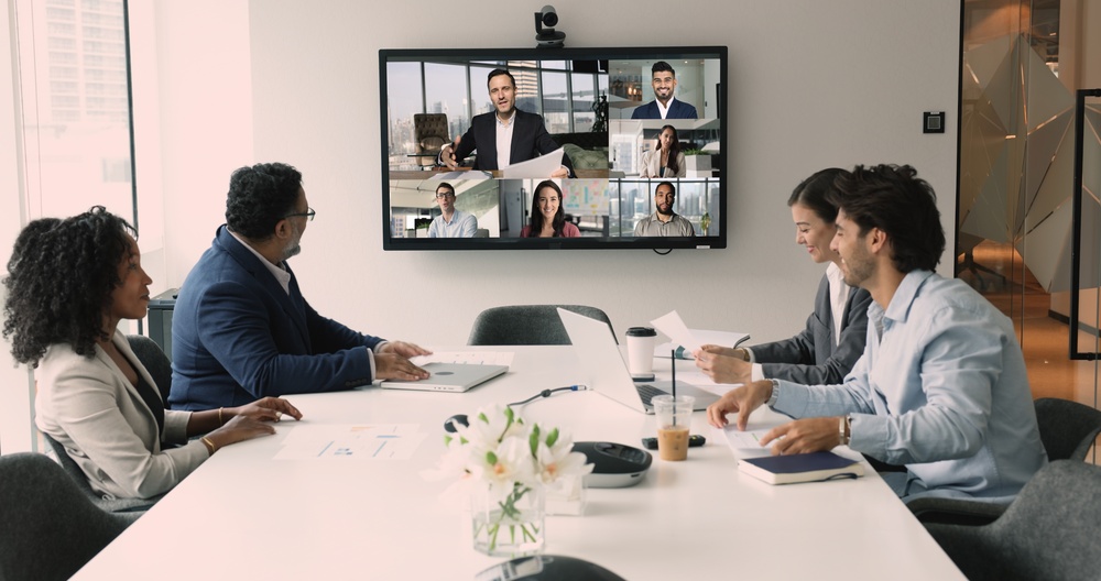 People sitting at a conference table in a video call meeting with remote participants shown on a wall screen