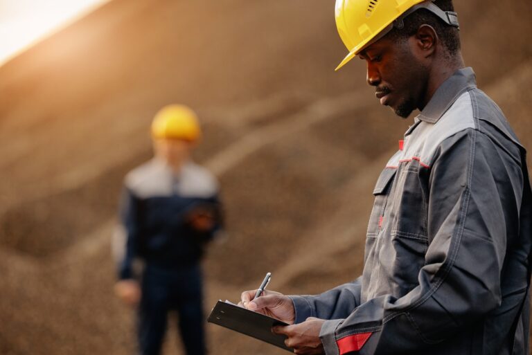 Man in a hard hat writing on a clipboard at a quarry, with another worker in the background
