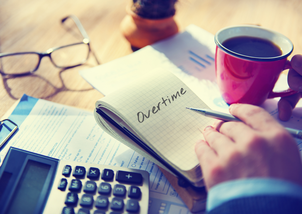 Person holding a coffee cup and pen over a notebook that reads Overtime, with a calculator, glasses, and paperwork nearby