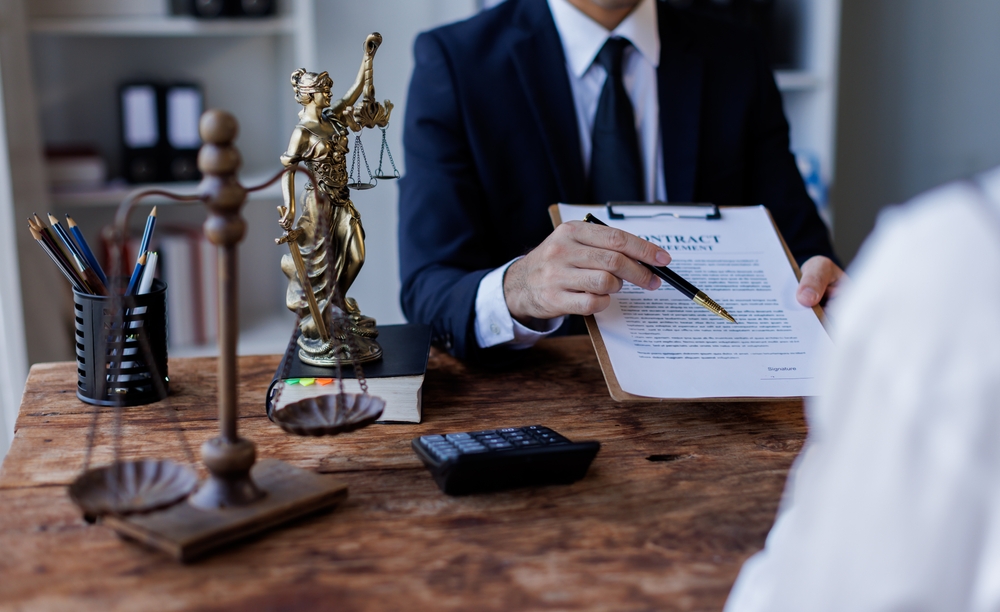 Man in a suit holding a clipboard with a document that reads Contract Agreement and Signature toward another person, with a statue of Lady Justice and a set of scales on the desk
