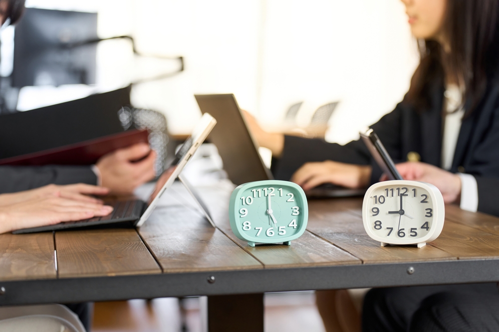 Two clocks on a table between people working on tablets and a laptop
