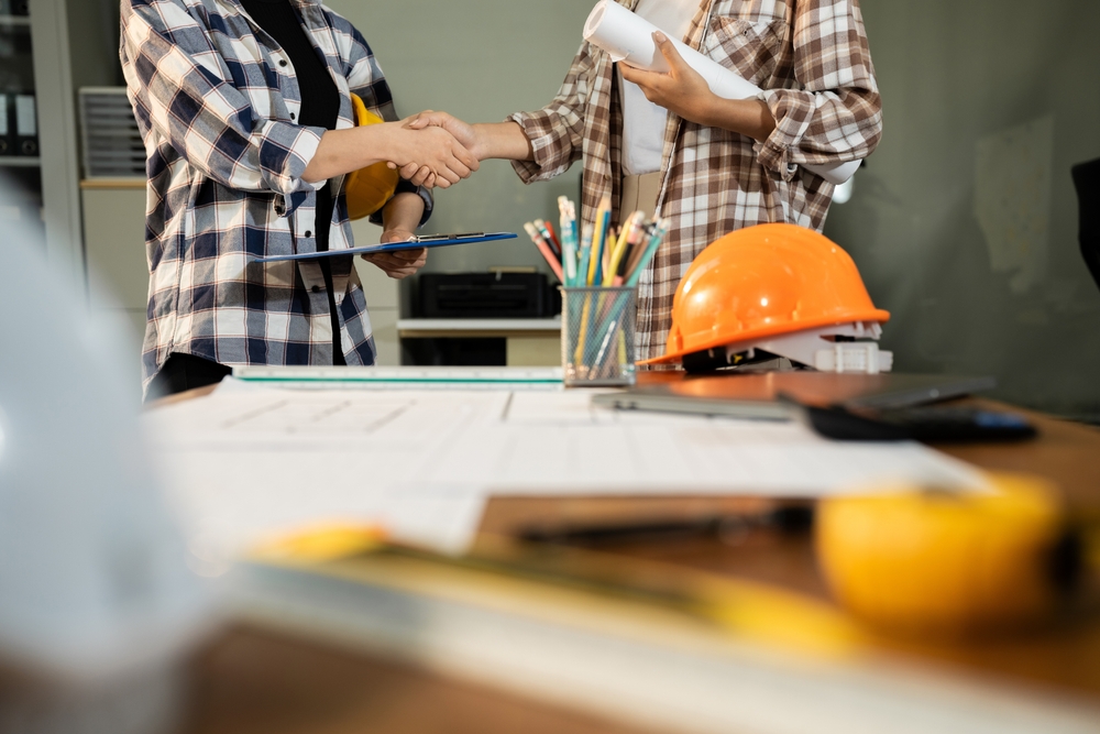 Two people shaking hands over a desk with blueprints and a hard hat, one holding a clipboard and the other holding rolled plans
